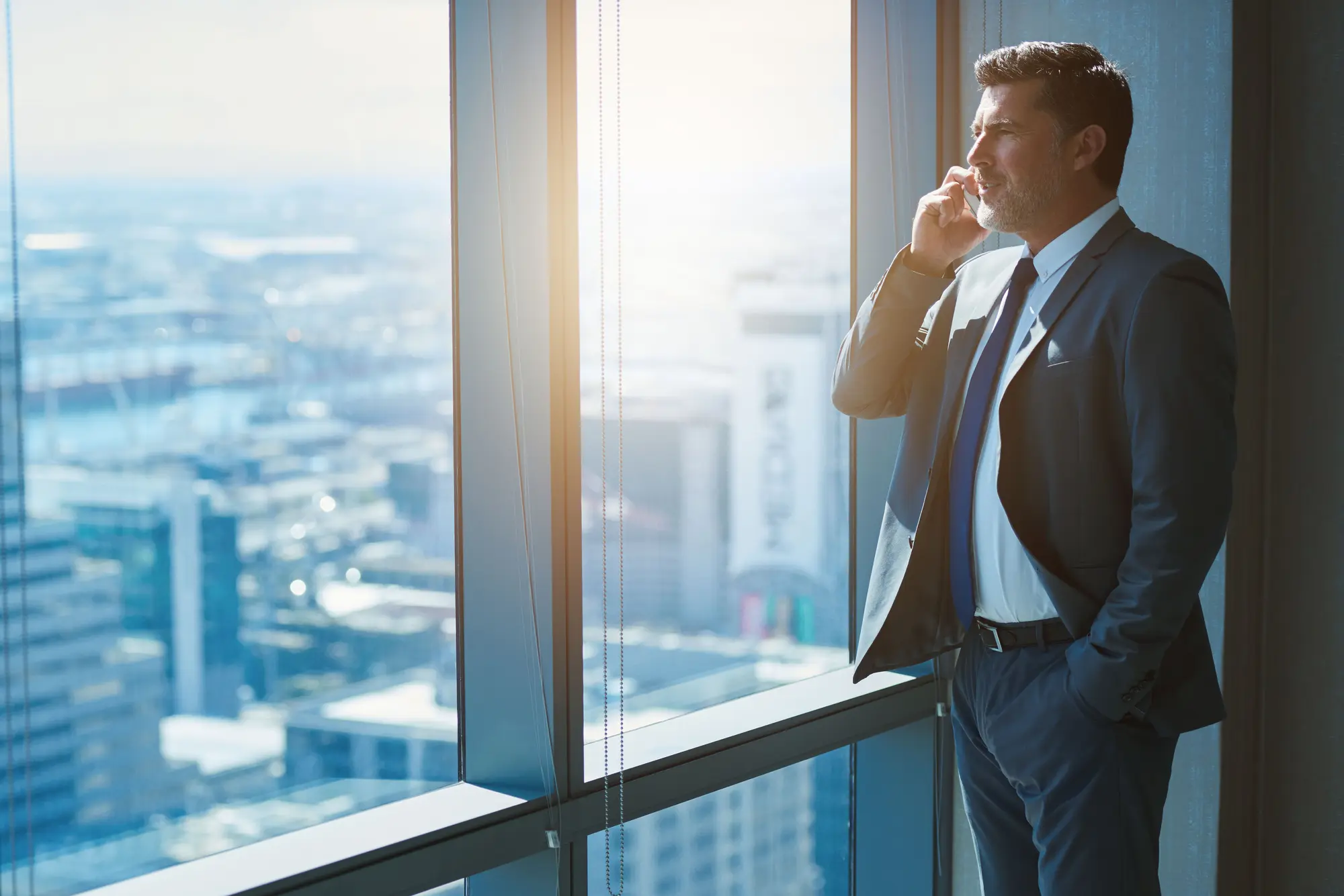 CPA NZ – Leading Certified Practising Accountant in New Zealand | Aurora Financials Handsome business CEO with designer stubble talking on phone in top-floor city office — representing a Certified Practising Accountant (CPA NZ) from Aurora Financials.