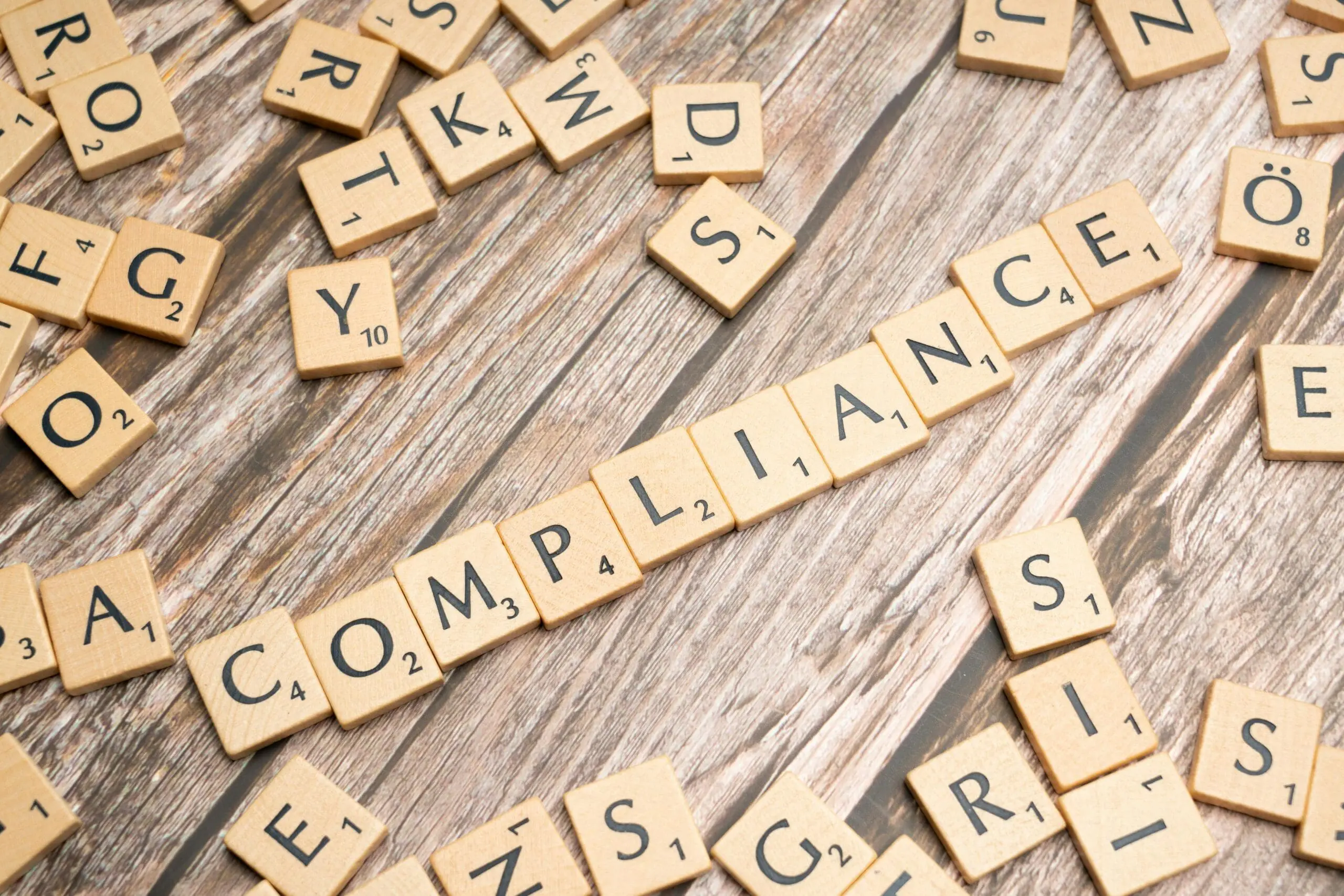 Wooden Scrabble tiles arranged on a wooden table spell out the word "COMPLIANCE," surrounded by scattered tiles with various letters.